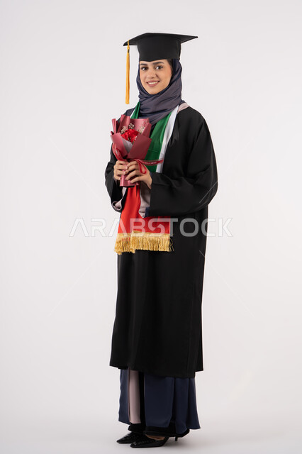 مناسبة واحتفال جامعي، الاحتفال بإنهاء الدراسة الجامعية، إيماءات السعادة بالوصول لحلم النجاح، بورتريه لخريجة عربية خليجية اماراتية محجبة ترتدي زي التخرج والقبعة تمسك بيدها بوكيه ورد تنظر إلى الكاميرا بتعابير السرور، الجسم بالطول الكامل، خلفية بيضاء