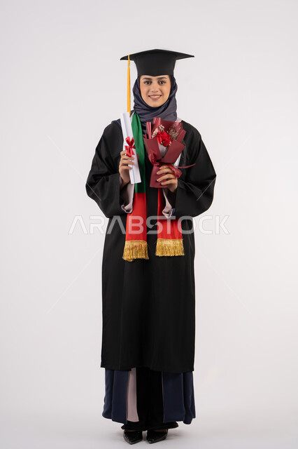 النظر إلى الكاميرا بتعابير السرور، مناسبة واحتفال جامعي، الاحتفال بإنهاء الدراسة الجامعية، إيماءات السعادة بالوصول لحلم النجاح، بورتريه لخريجة عربية خليجية اماراتية محجبة ترتدي زي التخرج والقبعة تمسك بيدها بوكيه ورد والشهادة، الجسم بالطول الكامل، خلفية بيضاء
