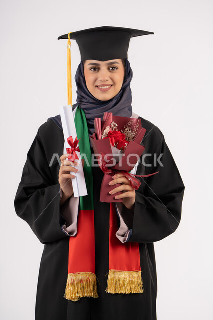 إيماءات السعادة بالوصول لحلم النجاح، مناسبة واحتفال جامعي، الاحتفال بإنهاء الدراسة الجامعية، مفهوم الاجتهاد والتفوق، النظر إلى الكاميرا بتعابير السرور، بورتريه لخريجة عربية خليجية اماراتية محجبة ترتدي زي التخرج والقبعة تمسك بيدها باقة ورد والشهادة، خلفية بيضاء