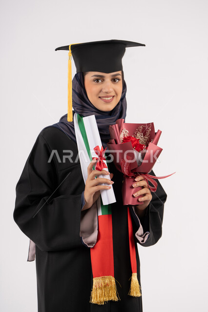 احتفال ومناسبة جامعية، الاحتفال بإنهاء الدراسة الجامعية، إيماءات السعادة بالوصول لحلم النجاح، مفهوم الاجتهاد والتفوق، النظر إلى الكاميرا بتعابير السرور، بورتريه لخريجة عربية خليجية اماراتية محجبة ترتدي زي التخرج والقبعة تمسك بيدها باقة ورد والشهادة، خلفية بيضاء