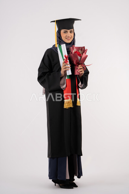 النظر إلى الكاميرا بتعابير السرور، مناسبة واحتفال جامعي، الاحتفال بإنهاء الدراسة الجامعية، إيماءات السعادة بالوصول لحلم النجاح، بورتريه لخريجة عربية خليجية اماراتية محجبة ترتدي زي التخرج والقبعة تمسك بيدها بوكيه ورد والشهادة، الجسم بالطول الكامل، خلفية بيضاء