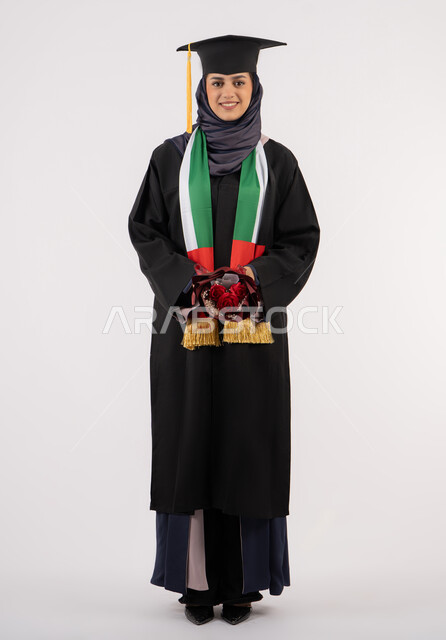 مناسبة واحتفال جامعي، الاحتفال بإنهاء الدراسة الجامعية، إيماءات السعادة بالوصول لحلم النجاح، بورتريه لخريجة عربية خليجية اماراتية محجبة ترتدي زي التخرج والقبعة تمسك بيدها بوكيه ورد تنظر إلى الكاميرا بتعابير السرور، الجسم بالطول الكامل، خلفية بيضاء