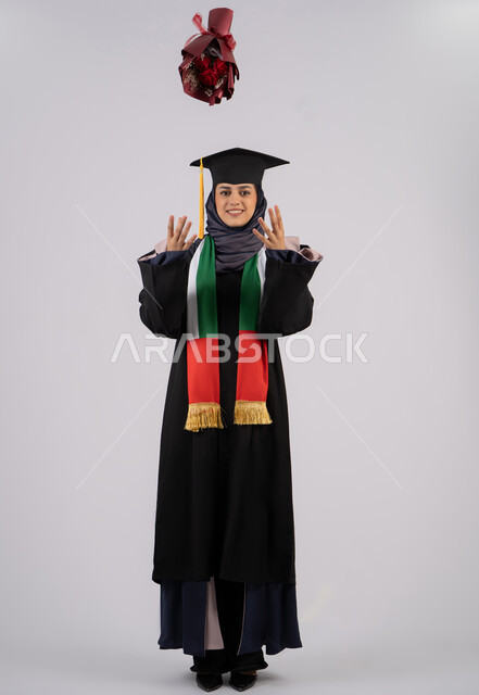 Throwing a bouquet of roses up with expressions of happiness and pleasure, university occasion and celebration, celebration of finishing university studies, portrait of a veiled Arab Gulf Emirati female graduate wearing graduation dress and cap looking at the camera with gestures of happiness for reaching the dream of success, full body, white background