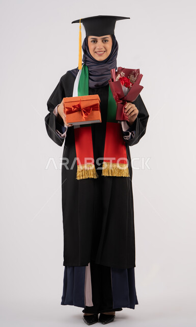 Celebrating the completion of university studies, giving gifts on occasions, university occasion and celebration, gestures of happiness for reaching the dream of success, portrait of a veiled Arab Gulf Emirati graduate wearing graduation dress and cap, holding a gift box with a bouquet of flowers, full body, white background