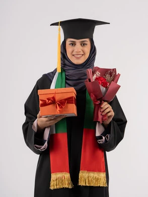 Giving gifts on happy occasions, university event and celebration, celebration of finishing university studies, gestures of happiness for reaching the dream of success, portrait of a veiled Arab Gulf Emirati graduate wearing graduation dress and cap holding a gift box with a bouquet of flowers, white background