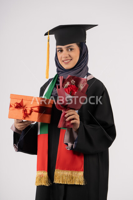 Expressions of happiness upon achieving the dream of success, celebrating the completion of university studies, giving gifts on occasions, university occasion and celebration, portrait of a veiled Arab Gulf Emirati graduate wearing a graduation gown and cap, holding a gift box with a bouquet of flowers, white background