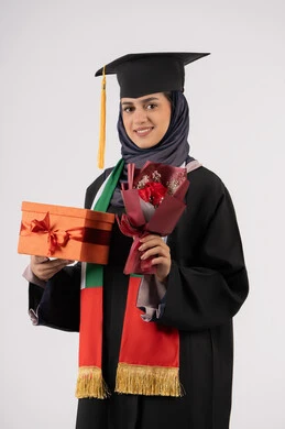 Expressions of happiness upon achieving the dream of success, celebrating the completion of university studies, giving gifts on occasions, university occasion and celebration, portrait of a veiled Arab Gulf Emirati graduate wearing a graduation gown and cap, holding a gift box with a bouquet of flowers, white background