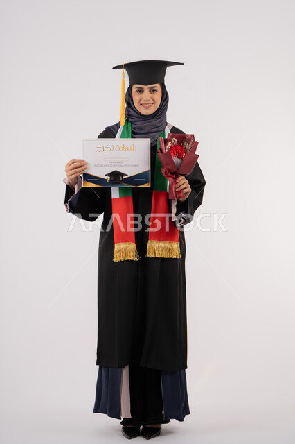 النظر إلى الكاميرا بتعابير السرور، مناسبة واحتفال جامعي، الاحتفال بإنهاء الدراسة الجامعية، إيماءات السعادة بالوصول لحلم النجاح، بورتريه لخريجة عربية خليجية اماراتية محجبة ترتدي زي التخرج والقبعة تمسك بيدها بوكيه ورد والشهادة، الجسم بالطول الكامل، خلفية بيضاء