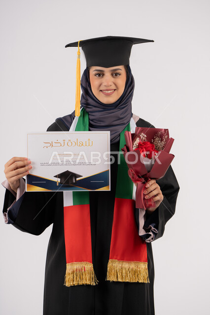 The concept of diligence and excellence, looking at the camera with expressions of pleasure, a university occasion and celebration, celebrating the completion of university studies, gestures of happiness for reaching the dream of success, a portrait of a veiled Arab Gulf Emirati graduate wearing a graduation gown and cap, holding a bouquet of flowers and a certificate, white background