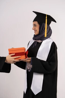 اهداء الهدايا بتعابير الفروح والسرور، بورتريه من الجانب لإمرأة عربية خليجية سعودية مبتسمة ترتدي قبعة وثوب التخرج تقوم بأخذ هدية بايماءات السعادة، خريجة اماراتية تحتفل بالنجاح وانهاء الدراسة الجامعية، خلفية بيضاء