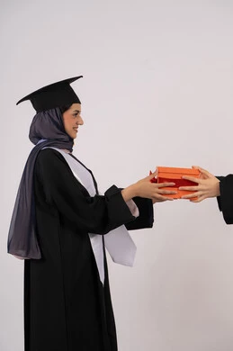 تقديم الهدايا بتعابير الفروح والسرور، بورتريه من الجانب لإمرأة عربية خليجية سعودية مبتسمة ترتدي قبعة وثوب التخرج تقوم بأخذ هدية بايماءات السعادة، خريجة اماراتية تحتفل بالنجاح وانهاء الدراسة الجامعية، خلفية بيضاء
