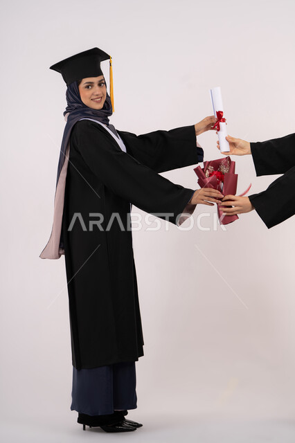 استلام الشهادة بتعابير الفروح والسرور، بورتريه من الجانب لإمرأة عربية خليجية سعودية ترتدي قبعة وثوب التخرج تقوم بأخذ باقة الورد تنظر الى الكاميرا بايماءات السعادة، خريجة اماراتية تحتفل بالنجاح وانهاء الدراسة الجامعية، صورة الجسم بالطول الكامل، خلفية بيضاء