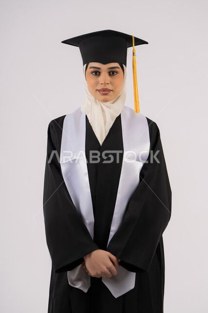 مفهوم التفوق والنجاح والاجتهاد، خريجة اماراتية تحتفل بإنهاء المرحلة الجامعية، الوقوف باستقامة والتقاط  صور شخصية بثياب التخرج، بورتريه لشابة عربية خليجية سعودية مبتسمة ترتدي القبعة وعباءة التخرج تنظر الى الكاميرا بتعابير السرور، خلفية بيضاء