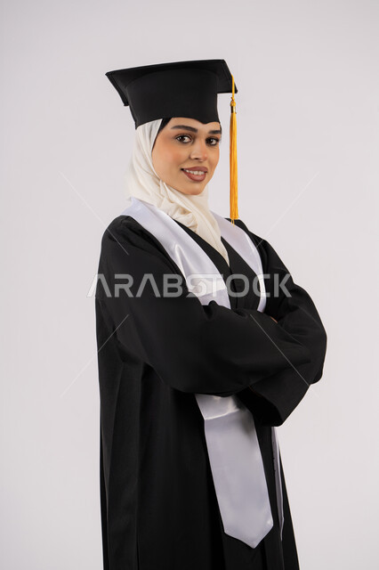 التقاط  صور شخصية بثياب التخرج، خريجة اماراتية تحتفل بإنهاء المرحلة الجامعية، بورتريه لشابة عربية خليجية سعودية مبتسمة ترتدي القبعة وعباءة التخرج تقف بأيدي متقاطعة وتنظر الى الكاميرا بتعابير السرور، مفهوم التفوق والنجاح والاجتهاد، خلفية بيضاء