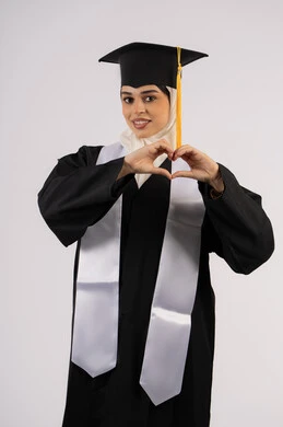 تشكيل اليدين على شكل قلب، التعبير عن الحب والفرح، التقاط  صور شخصية بثياب التخرج، خريجة اماراتية تحتفل بإنهاء المرحلة الجامعية، بورتريه لشابة عربية خليجية سعودية مبتسمة ترتدي القبعة وعباءة التخرج تنظر الى الكاميرا بتعابير السرور، خلفية بيضاء