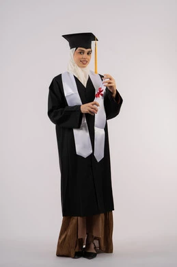 تعابير الانوثة واللطافة، التقاط  صور شخصية بثياب التخرج، خريجة اماراتية تحتفل بإنهاء المرحلة الجامعية، بورتريه لشابة عربية خليجية سعودية مبتسمة ترتدي القبعة وعباءة التخرج تحمل الشهادة وتنظر الى الكاميرا ايماءات السرور، صورة الجسم بالكامل، خلفية بيضاء