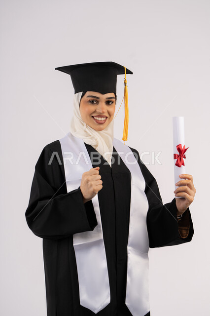 الفخر بالوصول إلى الأحلام، رفع قبضة اليد بتعابير الانجاز، النظر إلى الكاميرا بإيماءات السعادة والفرح، خريجة سعودية تحتفل بإنهاء المرحلة الجامعية، بورتريه لامرأة عربية خليجية اماراتية ترتدي القبعة وعباءة التخرج وتحمل الشهادة، خلفية بيضاء