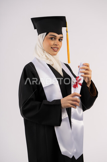 خريجة اماراتية تحتفل بإنهاء المرحلة الجامعية، تعابير الانوثة واللطافة، التقاط  صور شخصية بثياب التخرج، بورتريه لشابة عربية خليجية سعودية مبتسمة ترتدي القبعة وعباءة التخرج تحمل الشهادة وتنظر الى الكاميرا بإيماءات السرور والفرح، خلفية بيضاء