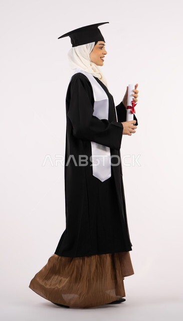 Celebrating reaching and achieving dreams, self-confidence gestures, taking selfies in graduation gowns, Saudi graduate celebrating finishing university, side portrait of an Arab Gulf Emirati woman wearing a graduation cap and gown and holding a diploma, full body photo, white background