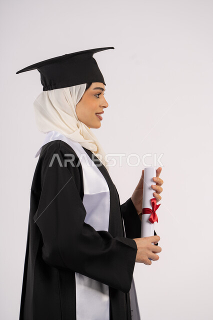 الوصول إلى الأحلام وتحقيقها، إيماءات الثقة بالنفس، التقاط  صور شخصية بثياب التخرج، خريجة سعودية تحتفل بإنهاء المرحلة الجامعية، بورتريه من الجانب لامرأة عربية خليجية اماراتية ترتدي القبعة وعباءة التخرج وتحمل الشهادة، خلفية بيضاء