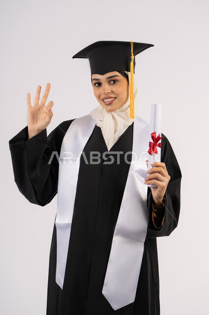 علامة الجودة والامتياز والتميز، الاحتفال بالوصول إلى الأحلام، خريجة سعودية تحتفل بإنهاء المرحلة الجامعية، بورتريه لامرأة عربية خليجية اماراتية ترتدي القبعة وعباءة التخرج وتحمل الشهادة تنظر إلى الكاميرا بإيماءات السعادة، خلفية بيضاء