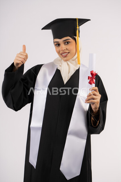 مناسبة وفرحة النجاح، رفع اصبع الابهام للأعلى بتعابير الاعجاب، التقاط  صور شخصية بثياب التخرج، خريجة اماراتية تحتفل بإنهاء المرحلة الجامعية، بورتريه لشابة عربية خليجية سعودية ترتدي القبعة وعباءة التخرج تحمل الشهادة وتنظر الى الكاميرا ايماءات السرور، خلفية بيضاء