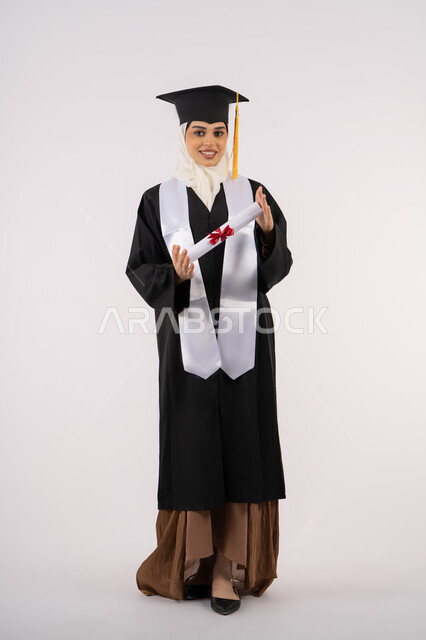 النظر إلى الكاميرا بإيماءات السعادة والفرح، الاحتفال بالوصول إلى الأحلام، التقاط  صور شخصية بثياب التخرج، خريجة سعودية تحتفل بإنهاء المرحلة الجامعية، بورتريه لامرأة عربية خليجية اماراتية ترتدي القبعة وعباءة التخرج وتحمل الشهادة، صورة الجسم بالكامل، خلفية بيضاء