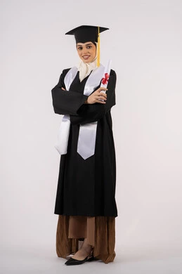 الوقوف بأيدي متقاطعة، الاحتفال بالوصول إلى الأحلام، خريجة سعودية تحتفل بإنهاء المرحلة الجامعية، بورتريه من  الجانب لامرأة عربية خليجية اماراتية ترتدي القبعة وعباءة التخرج وتحمل الشهادة تنظر إلى الكاميرا بإيماءات السعادة، صورة الجسم بالكامل، خلفية بيضاء