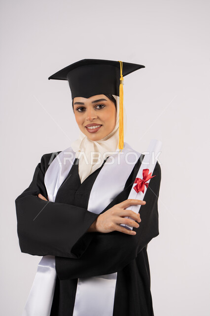 مفهوم النجاح والتفوق، الوقوف بأيدي متقاطعة، الاحتفال بالوصول إلى الأحلام، خريجة سعودية تحتفل بإنهاء المرحلة الجامعية، بورتريه من  الجانب لامرأة عربية خليجية اماراتية ترتدي القبعة وعباءة التخرج وتحمل الشهادة تنظر إلى الكاميرا بإيماءات السعادة، خلفية بيضاء