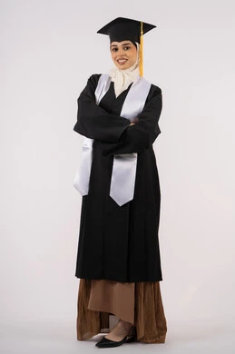 مفهوم الاجتهاد والتفوق، الوقوف بأيدي متقاطعة، الاحتفال بالوصول إلى الأحلام، خريجة سعودية تحتفل بإنهاء المرحلة الجامعية، بورتريه لشابة عربية خليجية اماراتية محجبة مبتسمة ترتدي ثياب التخرج تنظر إلى الكاميرا بإيماءات السرور والسعادة، صورة لكامل الجسم، خلفية بيضاء