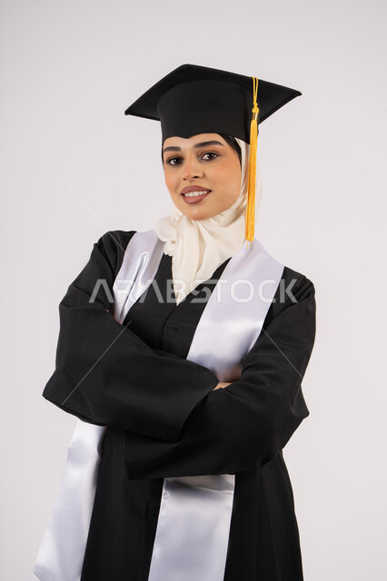 الاجتهاد والتفوق والنجاح، الوقوف بأيدي متقاطعة، الاحتفال بالوصول إلى الأحلام، خريجة سعودية تحتفل بإنهاء المرحلة الجامعية، بورتريه لشابة عربية خليجية اماراتية محجبة مبتسمة ترتدي ثياب التخرج تنظر إلى الكاميرا بإيماءات السرور والسعادة، خلفية بيضاء