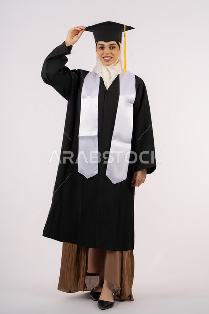 مفهوم الاجتهاد والتفوق، أهمية الدراسة الجامعية، الفرح بتحقيق النجاح, ايماءات البهجة والسعادة والفخر بالوصول لحلم التخرج، بورتريه لخريجة عربية سعودية خليجية محجبة مبتسمة ترتدي العباءة وقبعة التخرج، شابة إماراتية تحتفل بإنهاء دراستها، صورة لكامل الجسم، خلفية بيضاء