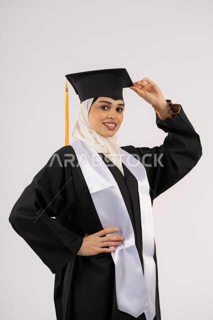 السرور والفرح بتحقيق النجاح, أهمية الدراسة الجامعية، مفهوم الاجتهاد والتفوق، ايماءات البهجة والسعادة والفخر بالوصول لحلم التخرج، بورتريه لخريجة عربية سعودية خليجية محجبة مبتسمة ترتدي العباءة وقبعة التخرج، شابة إماراتية تحتفل بإنهاء دراستها، خلفية بيضاء