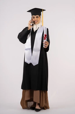 مفهوم الاجتهاد والتفوق، التواصل مع الأهل والأصدقاء، شابة سعودية تحتفل بإنهاء دراستها الجامعية، استخدام التقنية الحديثة،  بورتريه لخريجة عربية خليجية اماراتية ترتدي عباءه وقبعة التخرج تقوم بإجراء مكالمة عبر الهاتف المحمول، الجسم بالطول الكامل، خلفية بيضاء