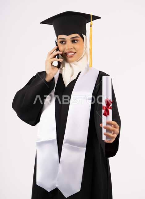التواصل مع الأهل والأصدقاء، شابة سعودية تحتفل بإنهاء دراستها الجامعية، استخدام التقنية الحديثة، الاحتفال بإنهاء الدراسة الجامعية، بورتريه لخريجة عربية خليجية اماراتية ترتدي عباءه وقبعة التخرج تقوم بإجراء مكالمة عبر الهاتف المحمول، خلفية بيضاء