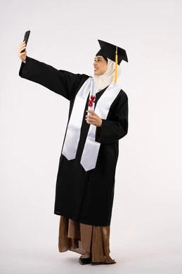 استخدام التقنية الحديثة، الاحتفاظ بالذكريات السعيدة، الاحتفال بإنهاء الدراسة الجامعية، بورتريه لخريجة عربية خليجية اماراتية ترتدي عباءه وقبعة التخرج تقوم بالتقاط صورة سيلفي تذكارية، الجسم بالطول الكامل، خلفية بيضاء