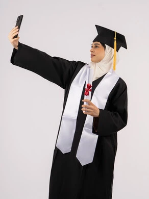الاحتفاظ بالذكريات السعيدة، استخدام التقنية الحديثة، الاحتفال بإنهاء الدراسة الجامعية، بورتريه لخريجة عربية خليجية اماراتية ترتدي عباءه وقبعة التخرج تقوم بالتقاط صورة سيلفي تذكارية، خلفية بيضاء