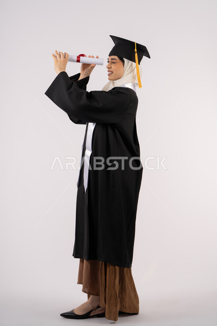 مشاعر الفخر والفرح بالوصول لحلم التخرج, التطلع إلى الأهداف والطموحات المستقبلية، بورتريه من الجانب لخريجة عربية سعودية خليجية محجبة ترتدي ثياب التخرج وتنظر من خلال شهادة التفوق، شابة إماراتية تحتفل بإنهاء دراستها، صورة لكامل الجسم، خلفية بيضاء