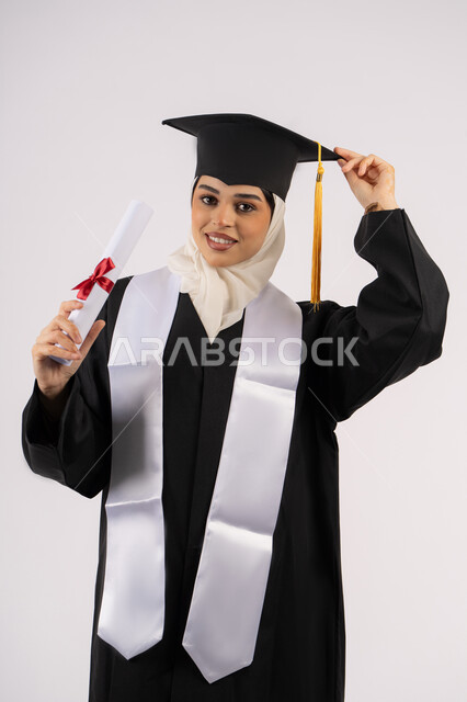 الفرح والفخر بالوصول لحلم التخرج, أهمية الدراسة الجامعية، بورتريه لخريجة عربية سعودية خليجية محجبة مبتسمة ترتدي ثياب التخرج وتحمل بيدها شهادة التفوق وتنظر إلى الكاميرا بإيماءات البهجة والسعادة، شابة إماراتية تحتفل بإنهاء دراستها، خلفية بيضاء