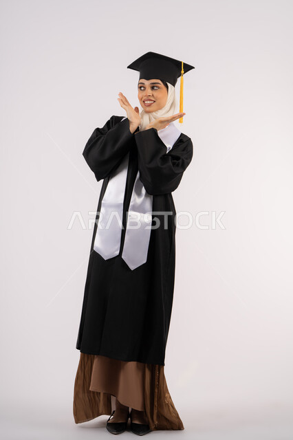 مفهوم التفوق والنجاح، تعابير الأنوثة واللطافة، التقاط  صور شخصية بثياب التخرج، خريجة اماراتية تحتفل بإنهاء المرحلة الجامعية، بورتريه لشابة عربية خليجية سعودية مبتسمة ترتدي القبعة وعباءة التخرج تقف بإيماءات السعادة والسرور، صورة الجسم بالكامل، خلفية بيضاء