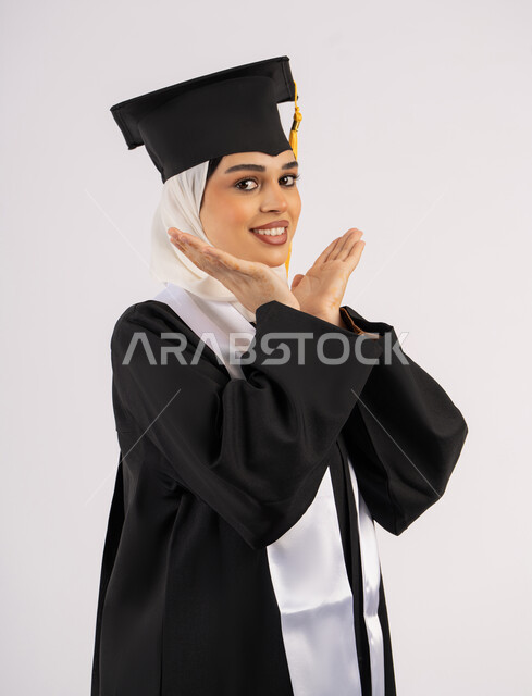 التقاط  صور شخصية بثياب التخرج، مفهوم التفوق والنجاح، تعابير الأنوثة واللطافة، خريجة اماراتية تحتفل بإنهاء المرحلة الجامعية، بورتريه لشابة عربية خليجية سعودية مبتسمة ترتدي القبعة وعباءة التخرج تقف بإيماءات السعادة والسرور، خلفية بيضاء
