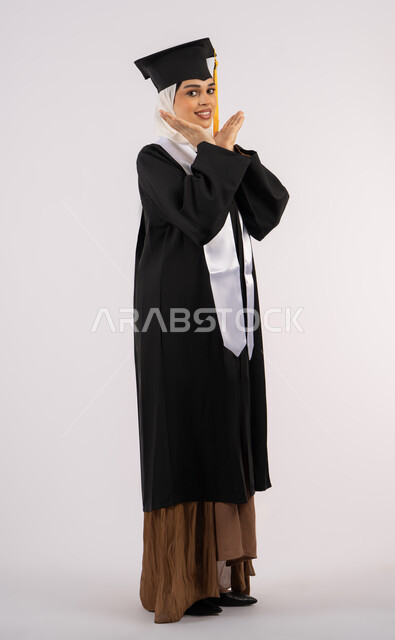 Expressions of femininity and tenderness, taking selfies in graduation gowns, Emirati graduate celebrating the completion of university, portrait of a smiling young Saudi Arabian Gulf woman wearing a graduation cap and gown looking at the camera with an expression of pleasure, concept of excellence and success, full body image, white background