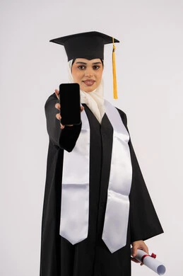 استخدام جهاز تقني حديث، عرض شاشة سوداء فارغة على الهاتف المحمول، تطبيقات الدراسة اونلاين، بورتريه لشابة عربية خليجية سعودية ترتدي القبعة وزي التخرج تمسك بيدها الشهادة، خريجة اماراتية مبتسمة تقف بتعابير السرور، الاحتفال بانتهاء المرحلة الجامعية، خلفية بيضاء