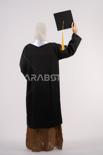 جودة التعليم في الامارات، الشعور بالفخر والفرح بالوصول لحلم التخرج, بورتريه من الخلف لخريجة عربية سعودية خليجية محجبة ترتدي عباءة وترفع قبعة التخرج، شابة إماراتية تحتفل بإنهاء دراستها الجامعية، صورة لكامل الجسم، خلفية بيضاء