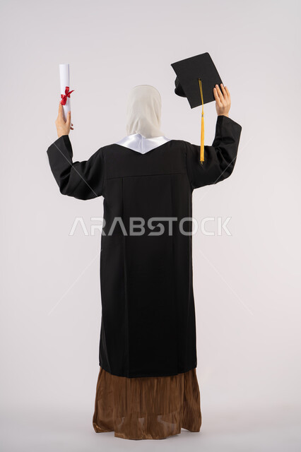 الشعور بالفخر والفرح بالوصول لحلم التخرج, جودة التعليم في الامارات، بورتريه من الخلف لخريجة عربية سعودية خليجية محجبة ترتدي عباءة وترفع قبعة التخرج وشهادة التفوق، شابة إماراتية تحتفل بإنهاء دراستها الجامعية، صورة لكامل الجسم، خلفية بيضاء