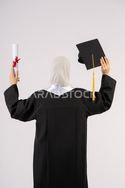 تحقيق النجاح والتفوق، الشعور بالفخر والفرح بالوصول لحلم التخرج, جودة التعليم في الامارات، بورتريه من الخلف لخريجة عربية سعودية خليجية محجبة ترتدي عباءة وترفع قبعة التخرج وشهادة التفوق، شابة إماراتية تحتفل بإنهاء دراستها الجامعية، خلفية بيضاء