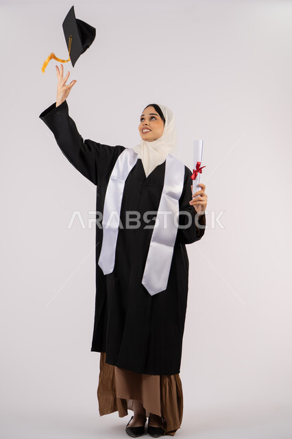 رمي القبعة احتفالا بالنجاح وانتهاء المرحلة الجامعية، بورتريه لخريجة عربية خليجية اماراتية ترتدي قبعة وزي التخرج تمسك بيدها الشهادة، شابة سعودية تقف بتعابير وايماءات السرور والسعادة، مفهوم التفوق والاجتهاد، صورة الجسم بالطول الكامل، خلفية بيضاء