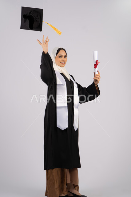 تحقيق التفوق والاجتهاد، رمي القبعة احتفالا بالنجاح وانتهاء المرحلة الجامعية، بورتريه لخريجة عربية خليجية اماراتية ترتدي قبعة وزي التخرج تمسك بيدها الشهادة، شابة سعودية تقف بتعابير وايماءات السرور والسعادة، صورة الجسم بالطول الكامل، خلفية بيضاء