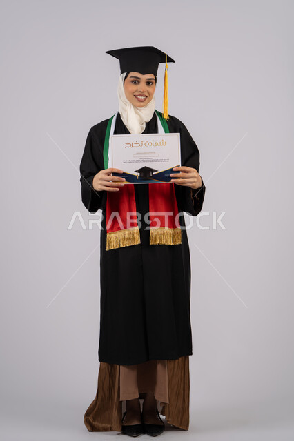 Celebrating the completion of university studies, looking at the camera with gestures of pride and happiness for achieving the dream, portrait of a veiled Arab Gulf Emirati graduate wearing a graduation gown and cap, holding a graduation certificate in her hand, full body, white background