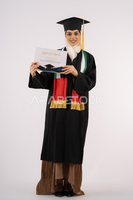 مناسبة واحتفال جامعي، النظر الى الكاميرا بايماءات الفخر والسعادة بالوصول الى الحلم، الاحتفال بإنهاء الدراسة الجامعية، بورتريه لخريجة عربية خليجية اماراتية محجبة ترتدي زي التخرج والقبعة تحمل بيدها شهادة التخرج، الجسم بالطول الكامل، خلفية بيضاء
